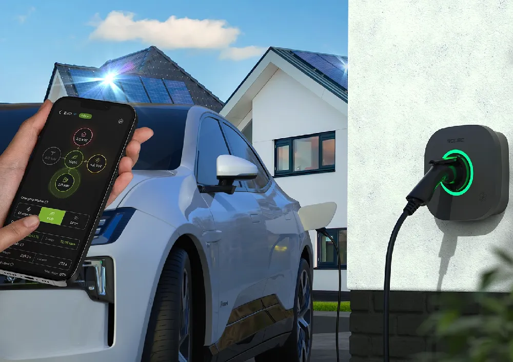 Hand holding a smartphone displaying the user friendly, easy to use Rolec EVO smart charging app in front of a white electric vehicle connected to a wall-mounted charger. A modern UK home with rooftop solar panels appears in the background.